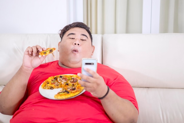 Uomo grasso che usa il suo smartphone mentre mastica una pizza