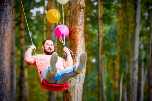 Uomo grasso e divertente su un'altalena con palloncini Ritorno all'infanzia