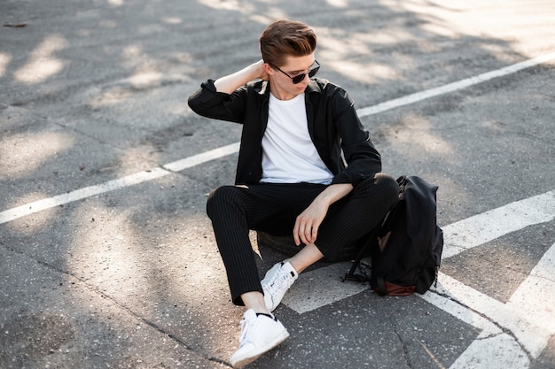 uomo hipster in vestiti neri in scarpe da ginnastica bianche alla moda con uno zaino in occhiali da sole si siede sulla strada.