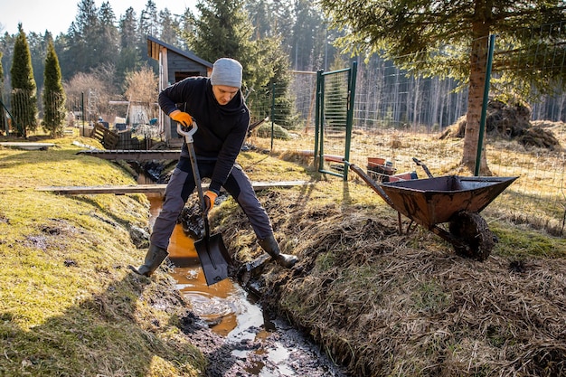 Uomo in abbigliamento da lavoro che pulisce un sentiero di giardino in piena acqua all'inizio della primavera con una pala e una carriola in mezzo a un paesaggio rurale