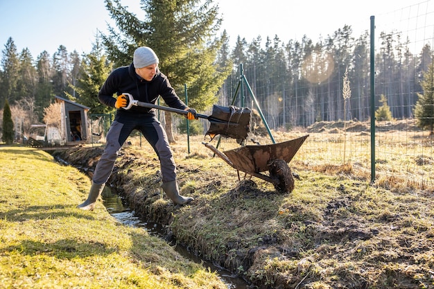 Uomo in abbigliamento da lavoro che pulisce un sentiero di giardino in piena acqua all'inizio della primavera con una pala e una carriola in mezzo a un paesaggio rurale