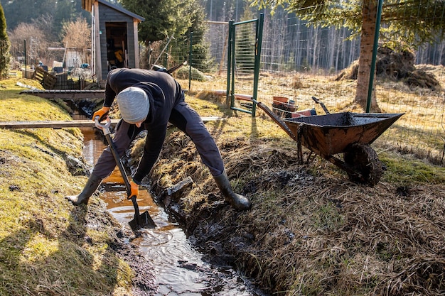 Uomo in abbigliamento da lavoro che pulisce un sentiero di giardino in piena acqua all'inizio della primavera con una pala e una carriola in mezzo a un paesaggio rurale