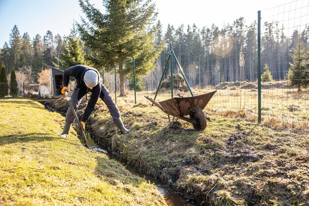 Uomo in abbigliamento da lavoro che pulisce un sentiero di giardino in piena acqua all'inizio della primavera con una pala e una carriola in mezzo a un paesaggio rurale