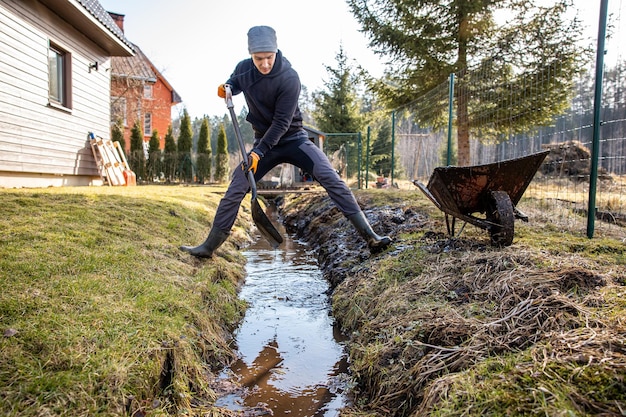 Uomo in abbigliamento da lavoro che pulisce un sentiero di giardino in piena acqua all'inizio della primavera con una pala e una carriola in mezzo a un paesaggio rurale