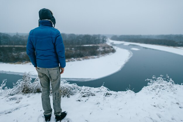 uomo in abiti caldi da dietro si erge sulla montagna innevata e guarda al fiume