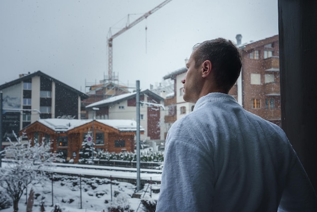 Uomo in accappatoio guarda fuori dalla finestra alla scena di zermatt innevata