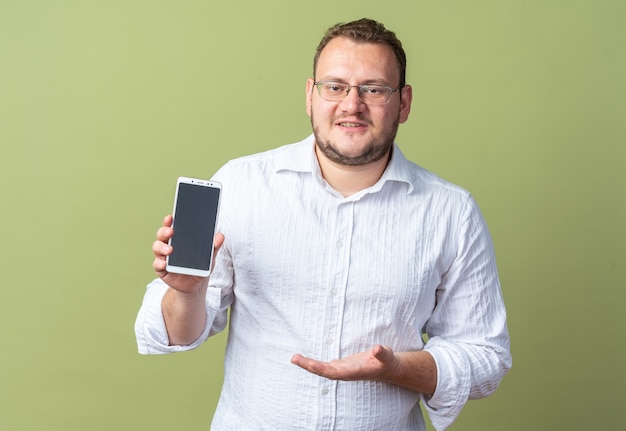 Uomo in camicia bianca con gli occhiali che mostra lo smartphone che lo presenta con il braccio della mano sorridente fiducioso in piedi sul muro verde