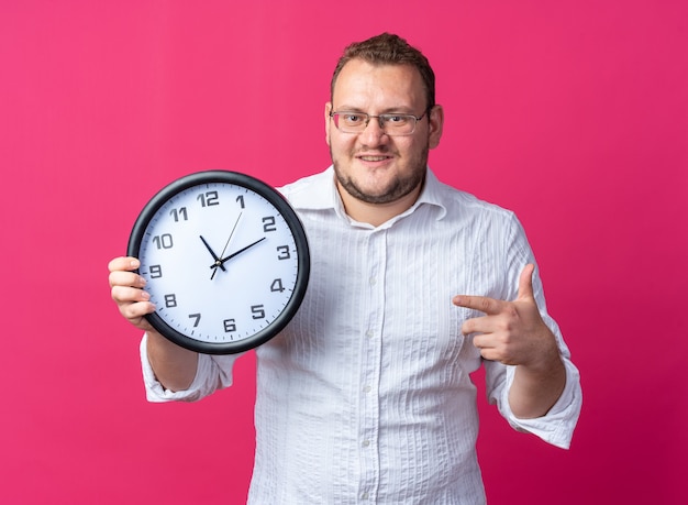 Uomo in camicia bianca con gli occhiali che tiene l'orologio da parete che punta con il dito indice e sembra sorridente felice e allegro