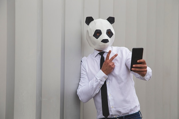 Uomo in camicia bianca, cravatta e maschera da panda che si fa selfie con lo smartphone contro il muro