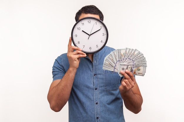 Uomo in camicia blu che si nasconde dietro l'orologio da parete che tiene in mano banconote da cento dollari in tempo per fare soldi Colpo di studio al coperto isolato su sfondo bianco