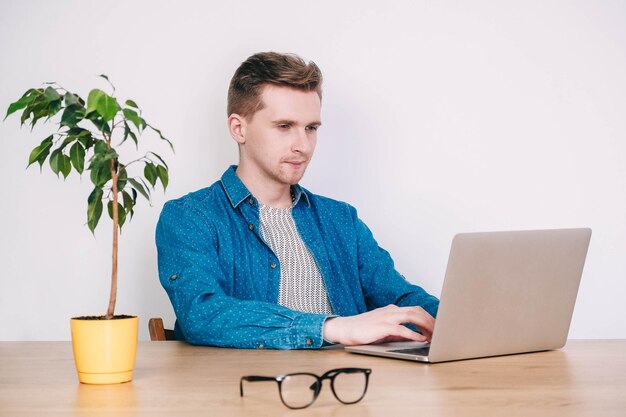 Uomo in camicia e occhiali che lavora al computer portatile seduto sul tavolo su uno sfondo bianco