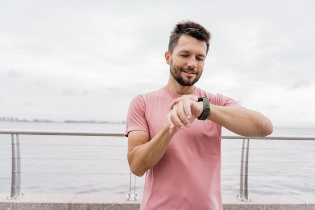 Uomo in camicia rosa che controlla il fitness tracker dopo l'allenamento contro un lungomare nuvoloso