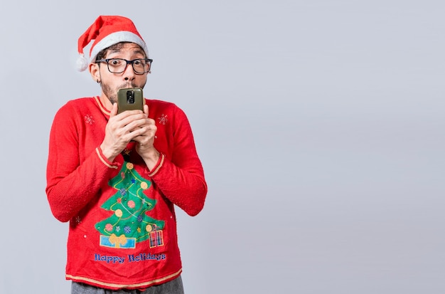 Uomo in cappello di Natale con il viso stupito che tiene il telefono Concetto di offerte speciali di Natale online Giovane uomo in abiti natalizi e cappello stupito tenendo il telefono