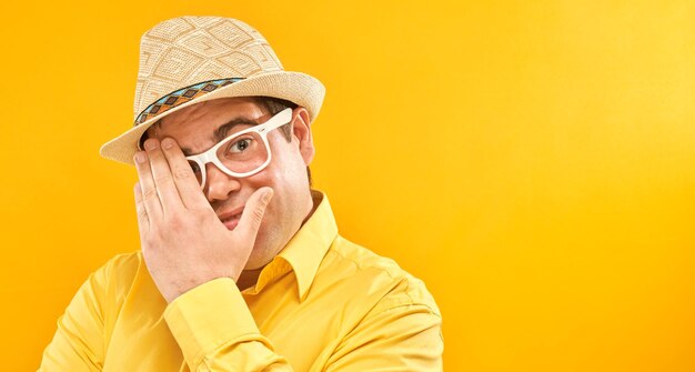 Uomo in cappello e occhiali che fa capolino attraverso le dita che coprono gli occhi con i palmi isolati su sfondo giallo Concetto di fobia