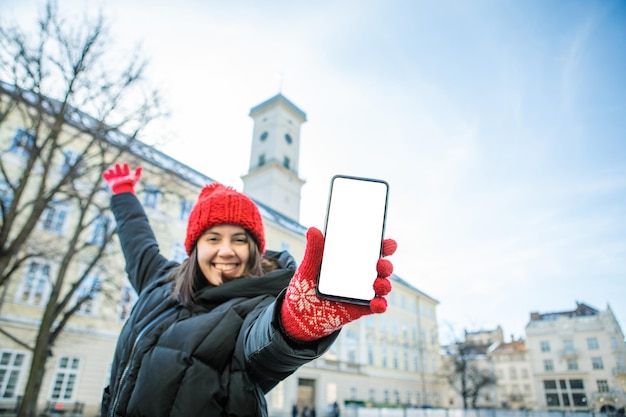 Uomo in cappello rosso invernale che tiene il telefono con il municipio europeo dello schermo vuoto bianco sullo sfondo