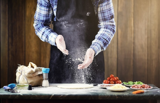 Uomo in grembiule che prepara una pizza, impasta l'impasto e mette gli ingredienti