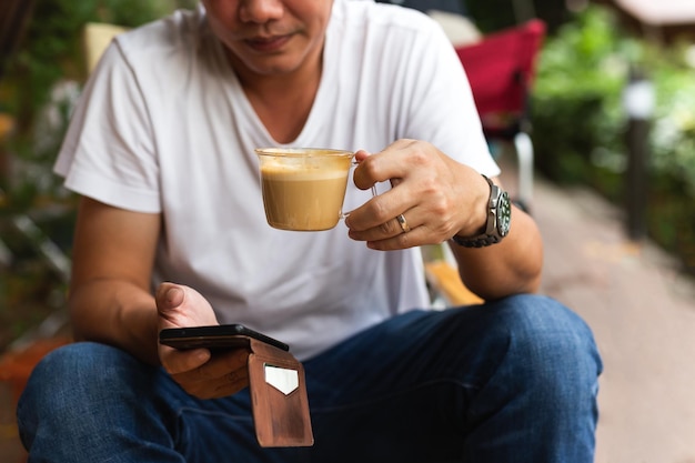 Uomo in jeans e maglietta bianca che beve caffè e guarda il cellulare nella caffetteria all'aperto