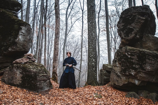 Uomo in kimono nero che pratica arti marziali con una spada su rocce e sullo sfondo della foresta