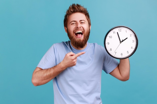 Uomo in maglietta blu che punta all'orologio da parete con il dito che guarda la fotocamera con un sorriso a trentadue denti che ride