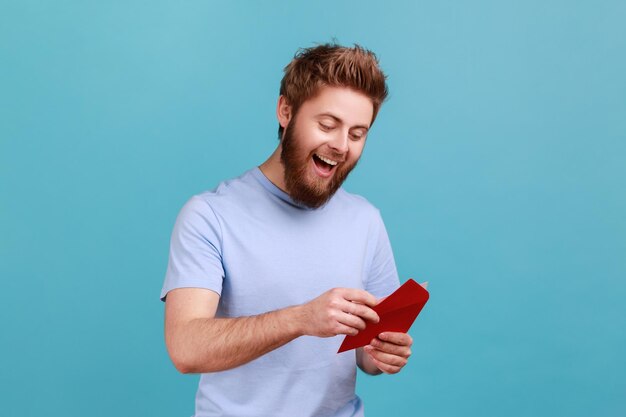 Uomo in maglietta che legge una lettera o un biglietto di auguri che tiene la busta sorridendo e rallegrandosi di notizie piacevoli