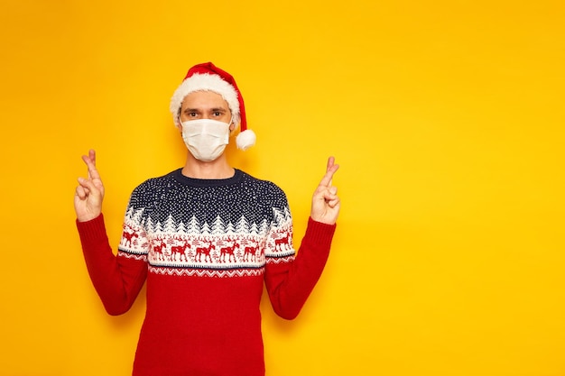 Uomo in maglione di capodanno cappello rosso di natale maschera medica isolato sfondo giallo dita incrociate