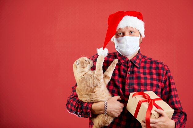 Uomo in maschera protettiva e cappello da Babbo Natale con regalo di Natale fai da te e un gatto