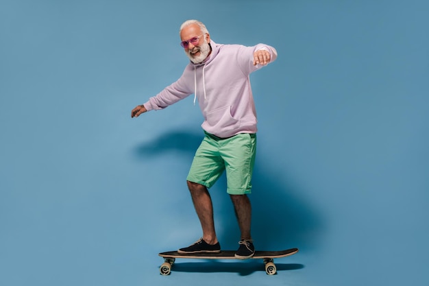 Uomo in occhiali da sole con cappuccio e pantaloncini in sella a skateboard Ragazzo adulto positivo con capelli bianchi in felpa lilla e camicia verde in posa