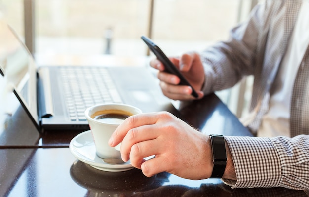 Uomo in orologio astuto che beve caffè sull'area di lavoro. Uomo che per mezzo del computer portatile, tenendo smart phone per lavoro aziendale o studio