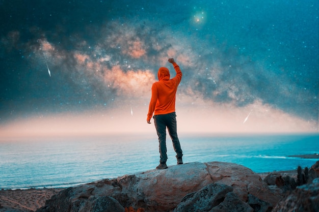 Uomo in piedi sulla cima della montagna vista posteriore con la mano alzata che fissa la Via Lattea e le stelle