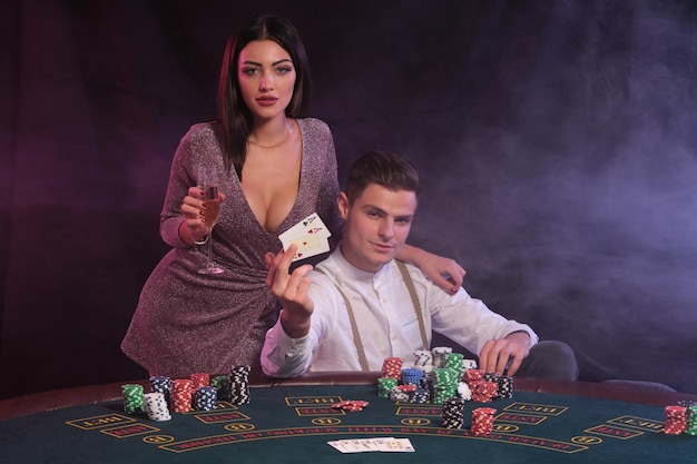 Uomo in possesso di carte giocare a poker al casinò seduto al tavolo con pile di fiches su di esso Celebra la vittoria con la donna Sfondo di fumo nero Closeup