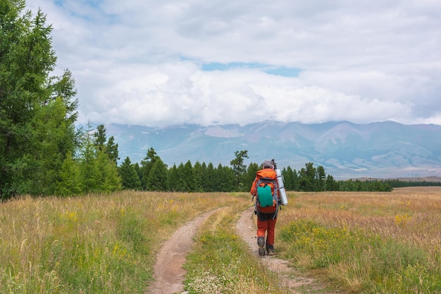 Uomo in rosso con un grande zaino in viaggio verso un'alta catena montuosa in nuvole basse Backpacker cammina attraverso la foresta verso grandi montagne sotto un cielo nuvoloso Paesaggio drammatico con turisti in montagna