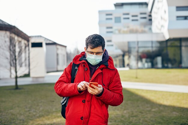 Uomo in rosso e con maschera di protezione usando il suo telefono per strada mentre covid19, ragazzo con gli occhiali con smartpone che cammina all'aperto in protezione per il viso