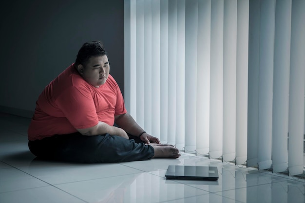Uomo in sovrappeso che guarda la bilancia vicino alla finestra