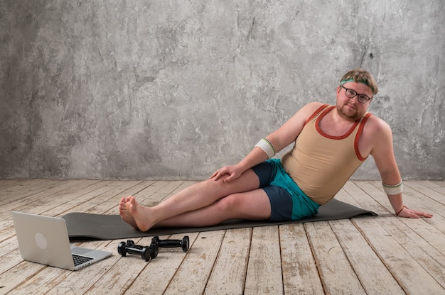 uomo in sovrappeso che si esercita, facendo esercizi di stretching sul tappetino yoga, guardando video di fitness online sul laptop a casa.