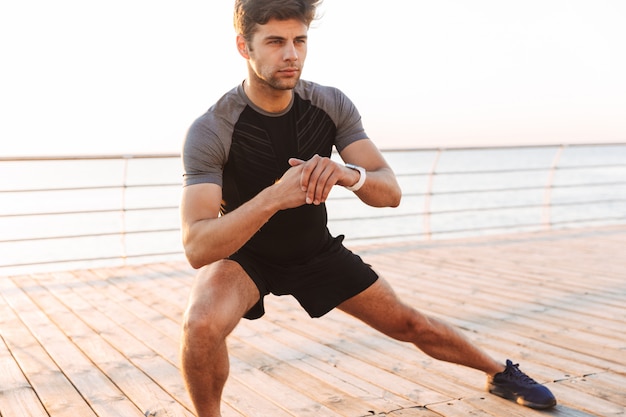 Uomo in tuta da ginnastica lavorando e facendo affondi laterali sul molo o sul lungomare in riva al mare, in mattinata