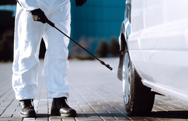 Uomo in tuta protettiva che lava e disinfetta le ruote dell'auto, per prevenire la diffusione del coronavirus.