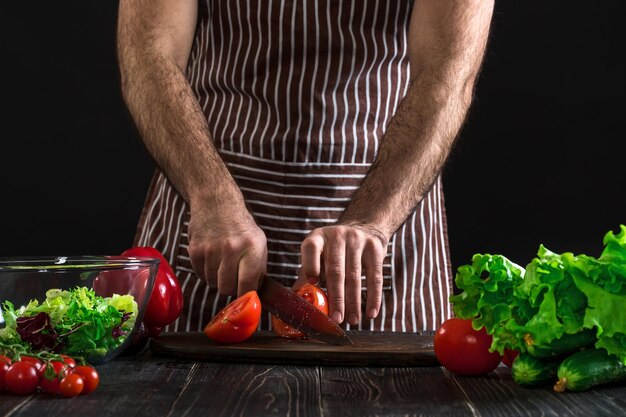 Uomo in un grembiule a righe che prepara insalata su un tavolo di legno. Le mani degli uomini tagliano il pomodoro per fare un'insalata su sfondo nero. Concetto di cibo sano