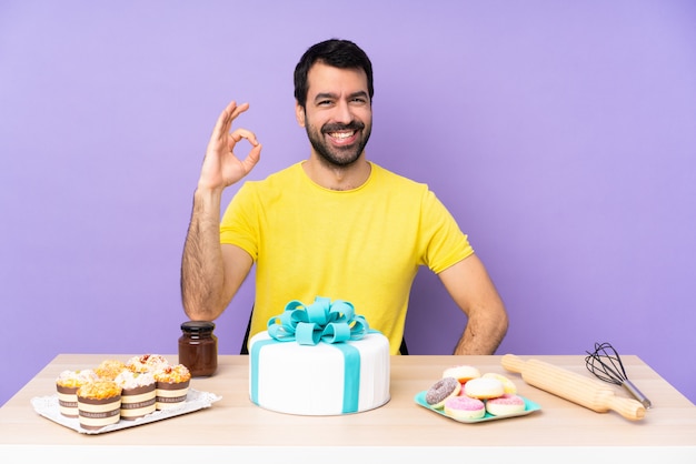 Uomo in una tabella con una grande torta che mostra segno giusto con le dita