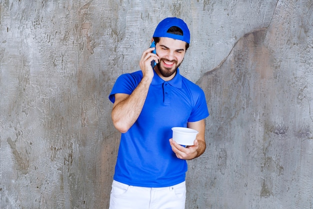 Uomo in uniforme blu che tiene un bicchiere di plastica da asporto e parla al telefono.