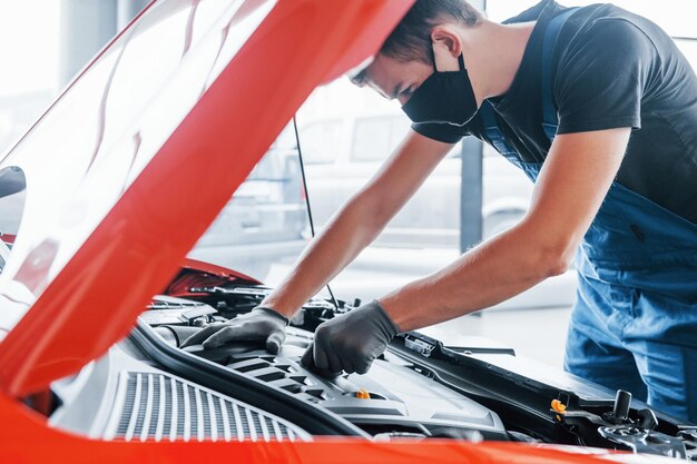 Uomo in uniforme e maschera protettiva nera lavora con automobile rotta Concezione del servizio auto