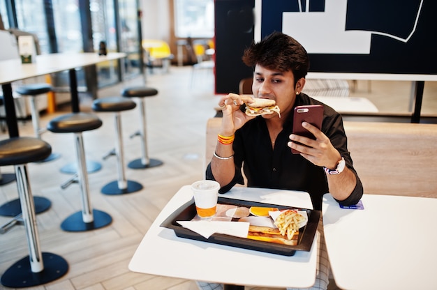 Uomo indiano alla moda e divertente che si siede al fast food e che mangia hamburger e che fa selfie dal telefono.