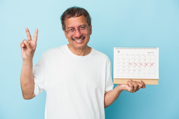Uomo indiano anziano che tiene un calendario isolato su sfondo blu gioioso e spensierato che mostra un simbolo di pace con le dita.
