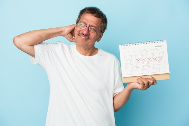 Uomo indiano anziano che tiene un calendario isolato su sfondo blu toccando la parte posteriore della testa, pensando e facendo una scelta.