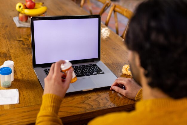 Uomo indiano con un farmaco in mano che fa una videochiamata usando un laptop, copia lo spazio sullo schermo a casa