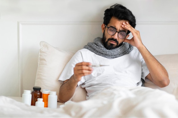 Uomo indiano malato che indossa una sciarpa seduto a letto e tiene in mano il termometro