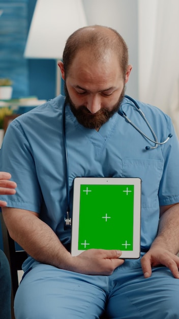 Uomo infermiere che tiene verticalmente lo schermo verde sul tablet mentre consulta il paziente con disabilità. Pensionato in sedia a rotelle che guarda la chiave cromatica con il modello di mockup isolato sul dispositivo.