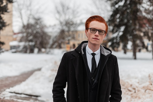 Uomo inglese bello ed elegante dai capelli rossi con gli occhiali e l'acconciatura in un abito alla moda con una cravatta e un cappotto nero in città. Stile di abbigliamento invernale da uomo