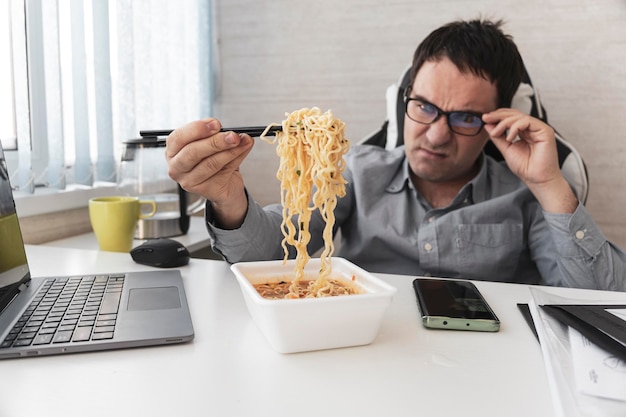 Uomo insoddisfatto che mangia spaghetti istantanei mentre lavora con il computer portatile in ufficio Pranzo in ufficio cibo spazzatura insapore