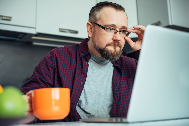 Uomo intelligente alla moda con la barba nell'interno moderno della cucina minimalista. Uomo d'affari in una camicia da casa facendo colazione e studiando le notizie guardando il laptop.