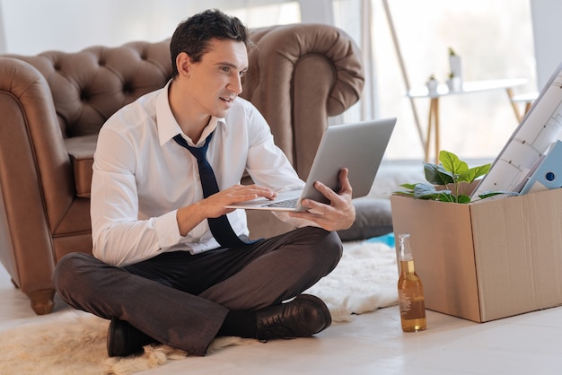 Uomo interessato. Calma giovane allegro guardando lo schermo del suo laptop mentre era seduto sul pavimento con le gambe incrociate mentre una bottiglia di alcol e una scatola di oggetti personali in piedi al suo fianco